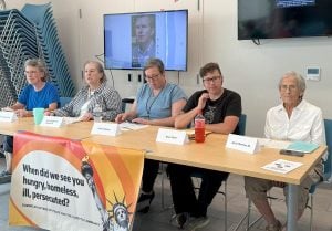 Five people sit in a pesentaion room behind a table with a name card in front of each person. A banner hanging from the front of the table shows the Statue of Liberty and reads "When did we see you hungry, homeless, ill, persecuted?"
