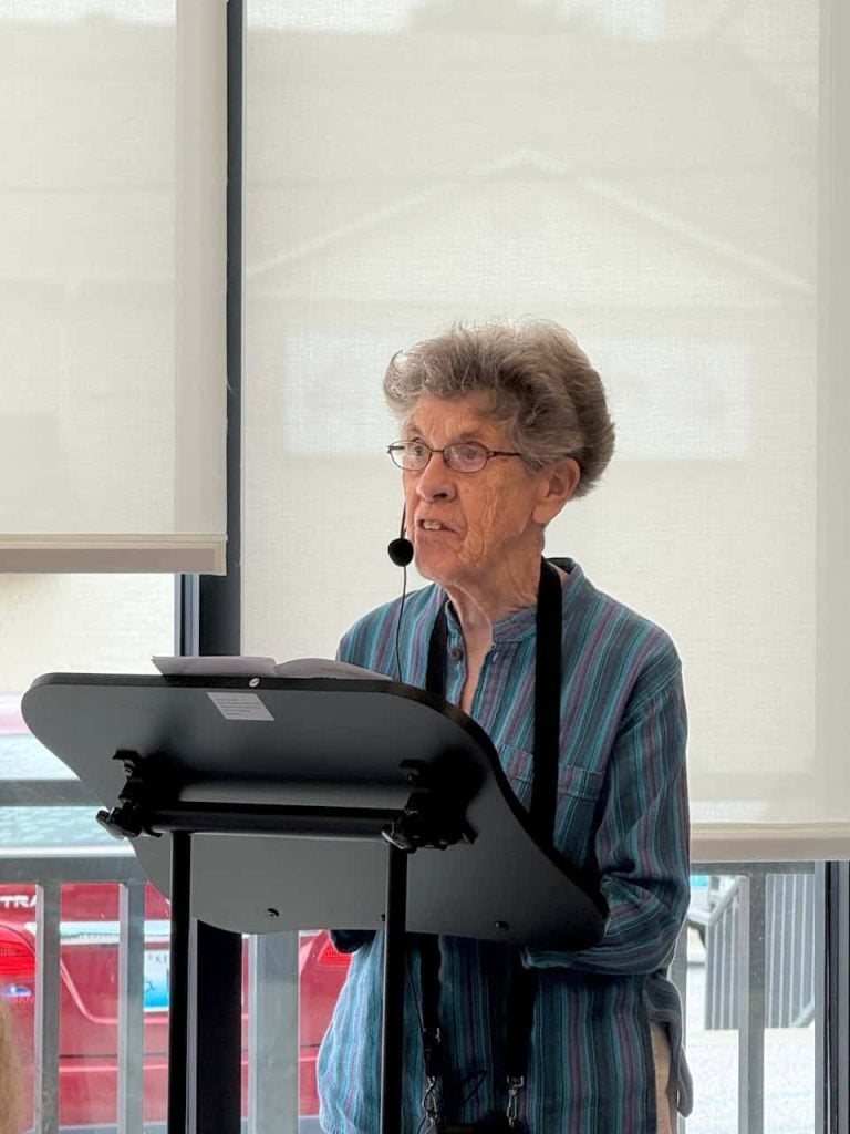 An older woman with short hair, round glasses, and a striped blue and purple shirt stands behind a podium and is speaking into a headset mic to an audience who isn't pictured.