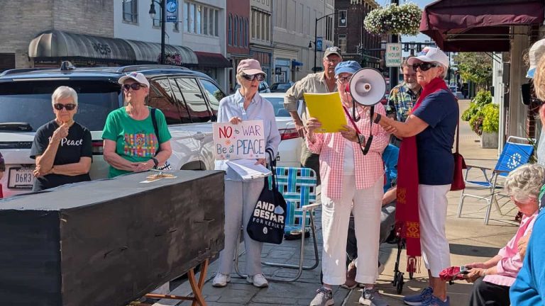 A group of older women with signs, banners and a megaphone demonstrate on the sidewalk.