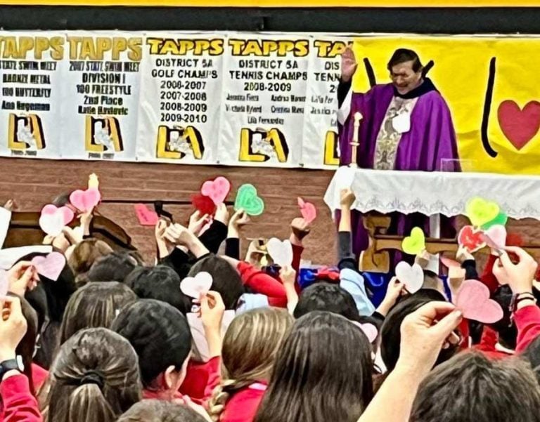 Children raise paper hearts over their heads during Catholic Mass in a school gym.