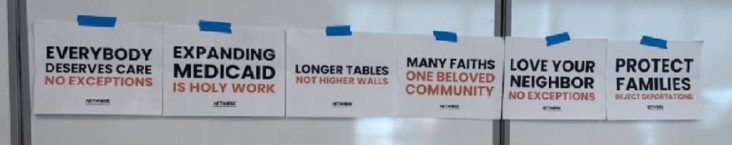 Posters that say "expanding medicaid is holy work" and "Longer tables not higher walls" "many faiths one beloved community" "love your neighbor no exceptions" "protect families reject deportations" "everybody deserves care no exceptions"