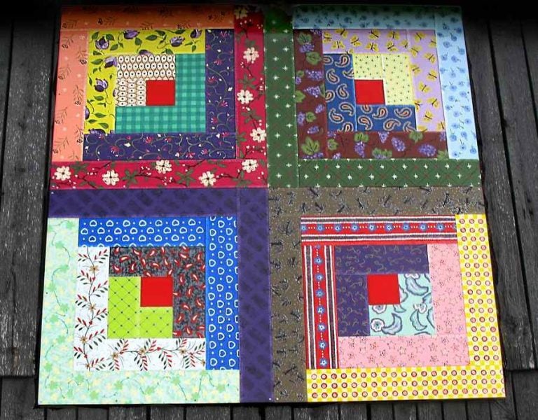 A quilt-like art piece hangs on the side of a wooden barn. The four quilt blocks are done in log cabin style, with a turkey red center square in each colorful quilt block.