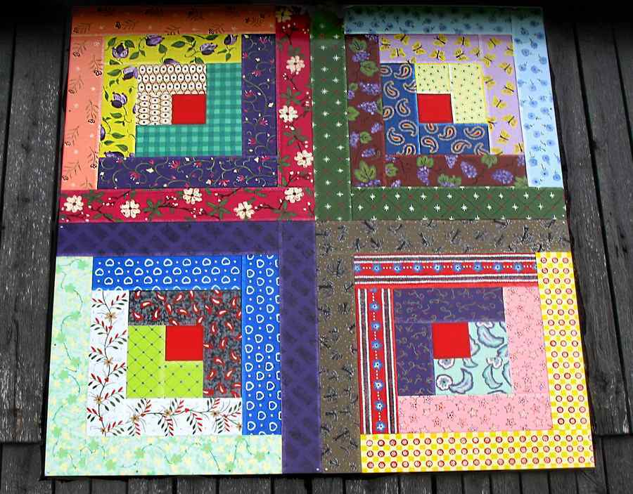 A quilt-like art piece hangs on the side of a wooden barn. The four quilt blocks are done in log cabin style, with a turkey red center square in each colorful quilt block.
