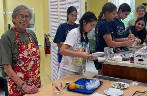 A couple older women are pictured helping high school students make scones in a kitchen.