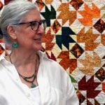 An older white woman with grey hair in a bun and a white shirt and turquoise jewelry with black glasses stands in front of a orange pattered quilt.