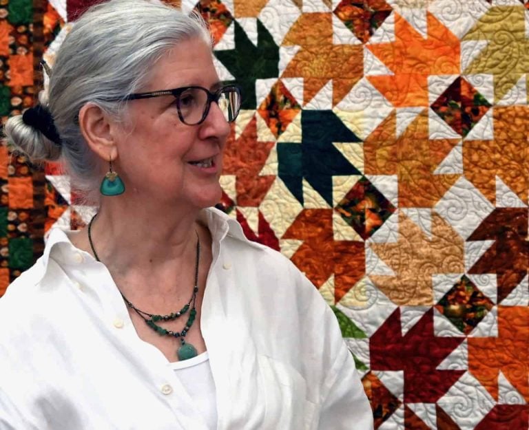 An older white woman with grey hair in a bun and a white shirt and turquoise jewelry with black glasses stands in front of a orange pattered quilt.
