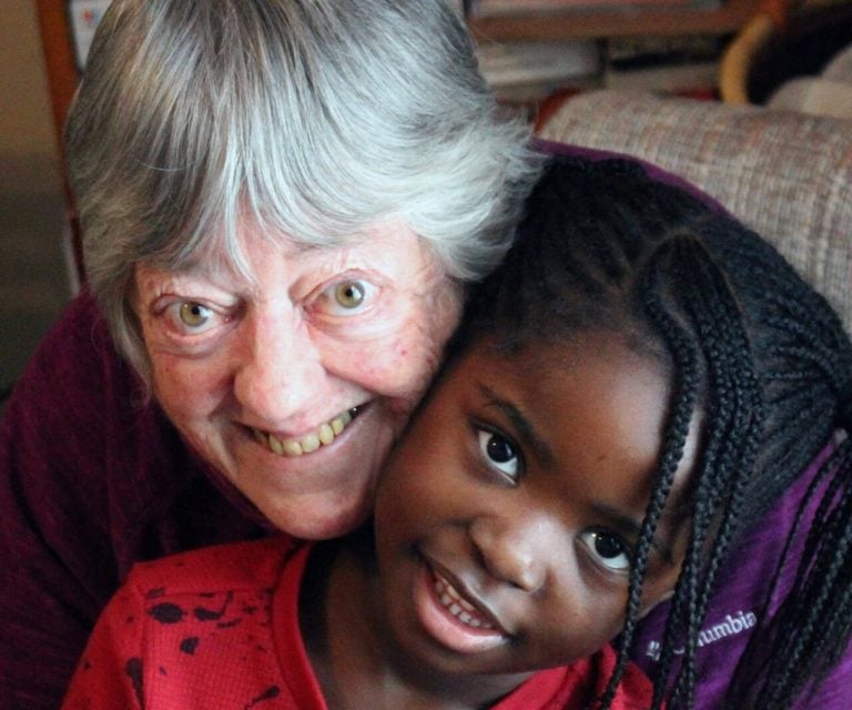 An older white woman with grey hair smiles with cheeks pressed together with a young Black girl with long cornrows in two pigtails.