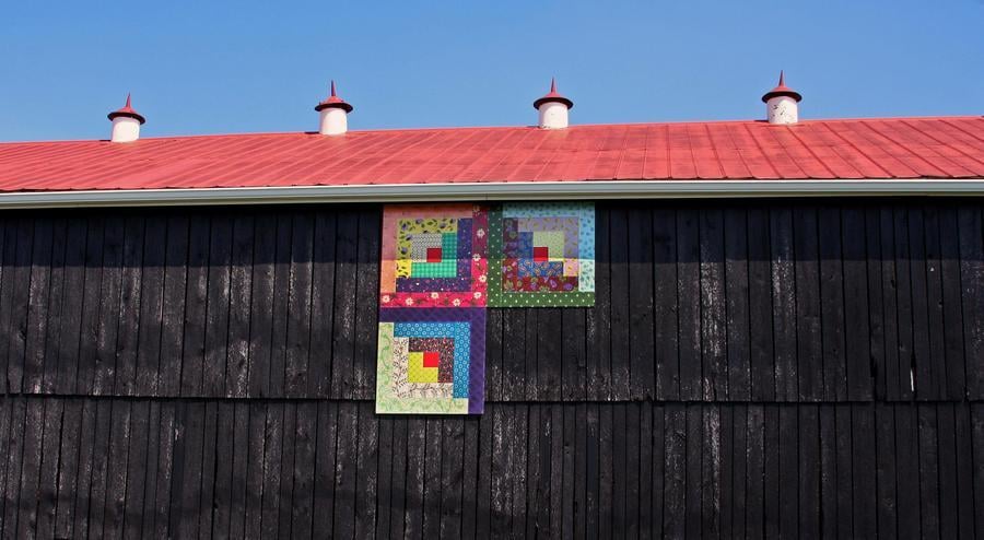 Three of four colorful painted quilt blocks hang on the side of an old wooden barn.