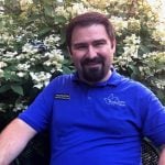 A white man sitting down outside smiling. He has short brown hair and a brown goatee and is wearing a bright blue collared shirt with the words "Loretto Motherhouse" on it.