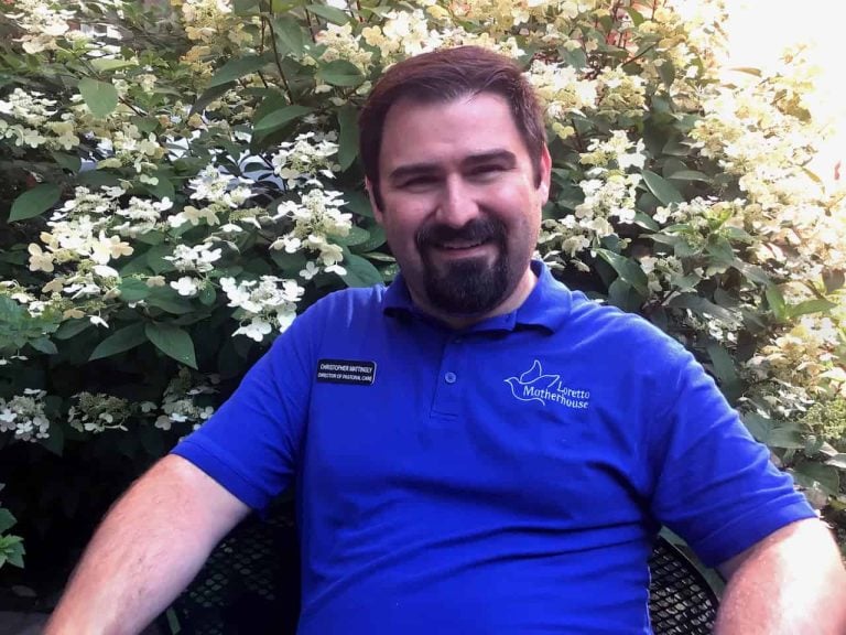 A white man sitting down outside smiling. He has short brown hair and a brown goatee and is wearing a bright blue collared shirt with the words "Loretto Motherhouse" on it.