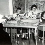 A black and white photo of students using headphones and recording equipment as a nun is reading to them.
