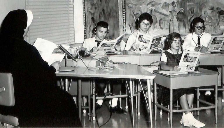 A black and white photo of students using headphones and recording equipment as a nun is reading to them.