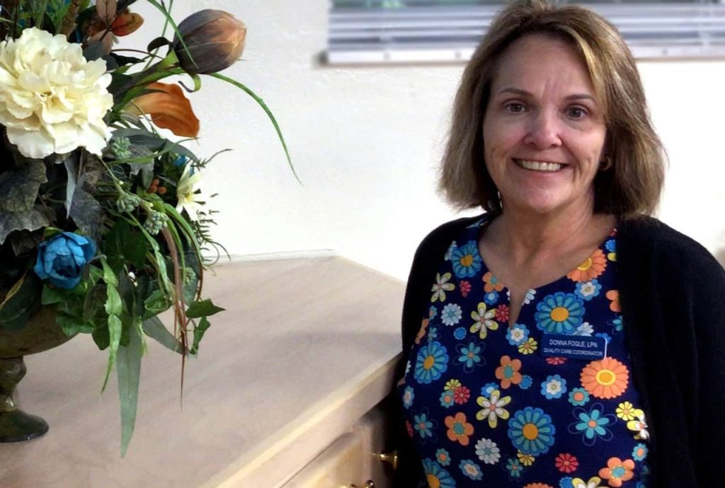 A woman with brightly colored flowers scrubs and shoulder length light brown hair smiles next to a vase of flowers.