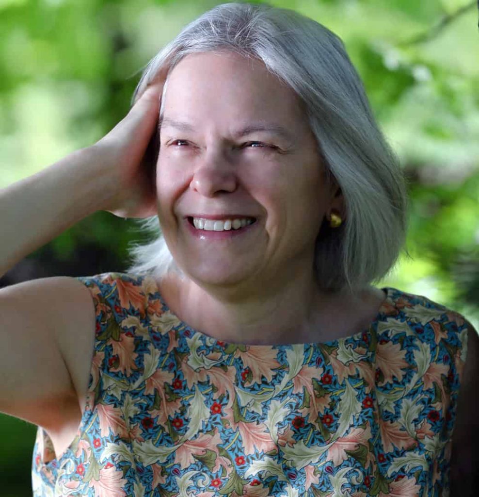 A middle aged white woman with short grey hair, gold hoop earrings a colorful shirt with blue, red, orange and green.