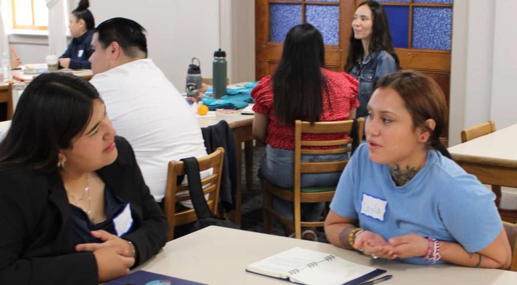 A group of college Latino students sitting at tables during an orientation and talking to one another.