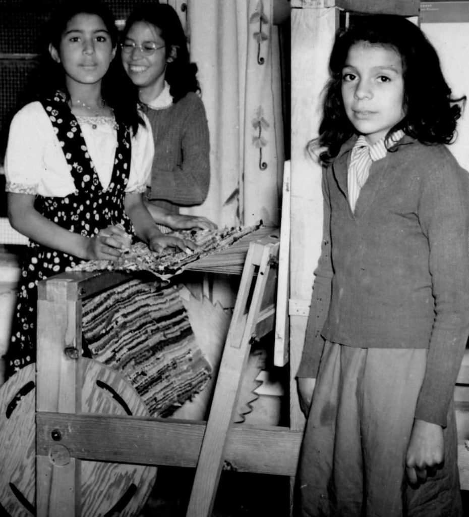 A black and white photo of three school girls weaving on a loom.