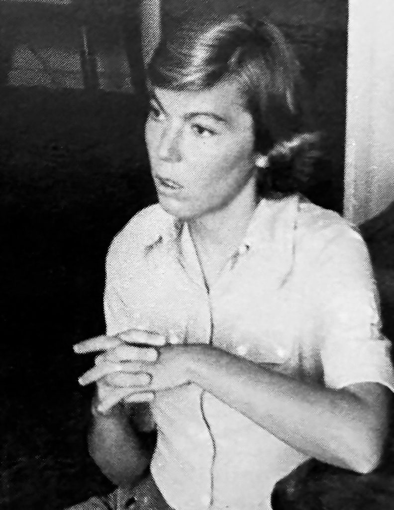 A black and white photo of a young woman looking away from the camera with her hands clasped together talking.