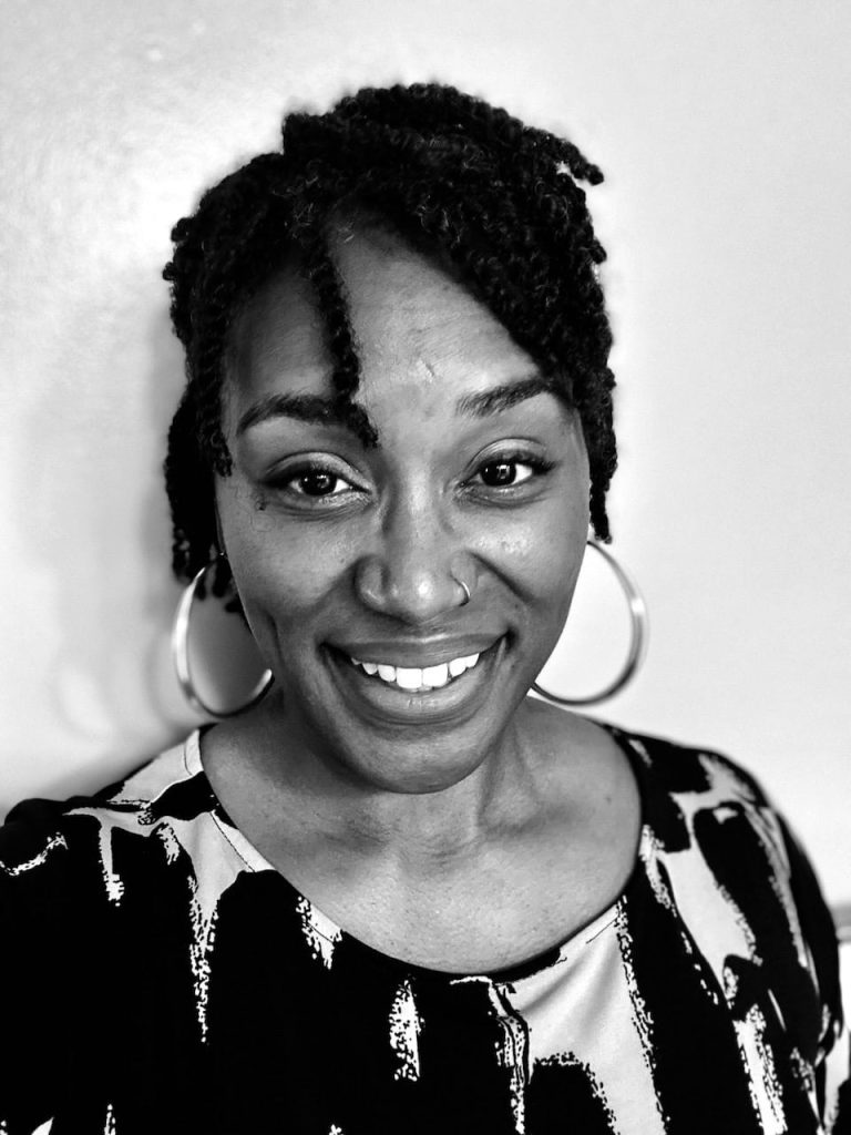 A black and white photo of a middle aged Black woman with her hair up in twists smiling wearing big hoop earrings and a nose ring.