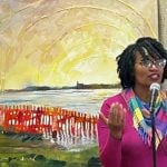 A black woman with natural hair, a pink shirt and glasses stands in front of her mixed media piece she made during an artist residency. The piece has green grass, a red fence, an ocean and a bright yellow sky.