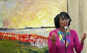 A black woman with natural hair, a pink shirt and glasses stands in front of her mixed media piece she made during an artist residency. The piece has green grass, a red fence, an ocean and a bright yellow sky.