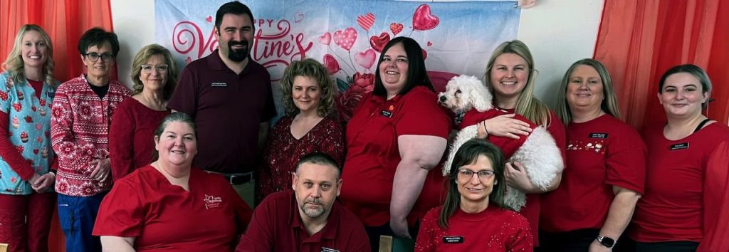 Dedicated and skilled Loretto Living Center staff members take a moment from their busy day for a photo. They are all dressed in red for valentines day and one woman is holding a fluffy white dog that is wearing a red shirt