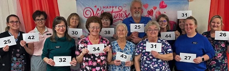 A group of nursing home staff members on valentines day holding up signs with numbers that signify how long they have worked at the Loretto Living Center. The signs range from 25-44 years.