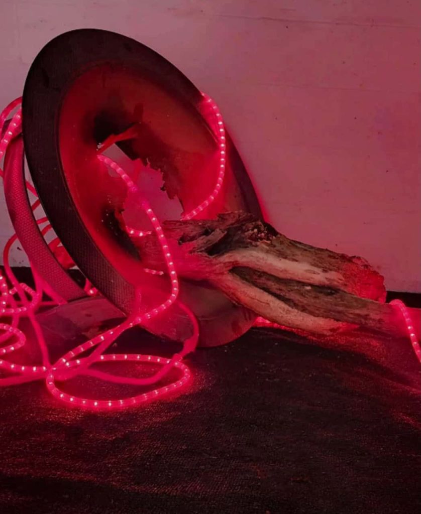 An art piece with a circle of metal with a large branch coming through the middle wrapped in red lights.