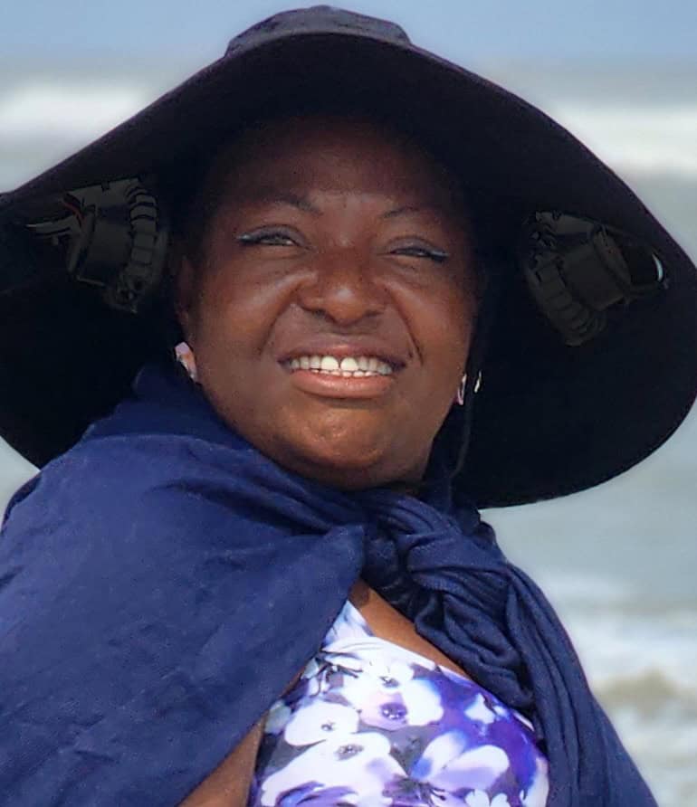 A black woman smiling. She wears a hat with two fans in the brim and is wearing a blue shawl.