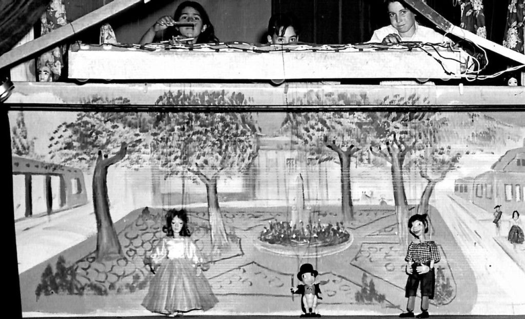 A black and white photo of children standing above a cardboard set as they give a marionette show.
