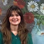 A woman with long brown hair and wispy bangs smiles in front of a painted flower mural.