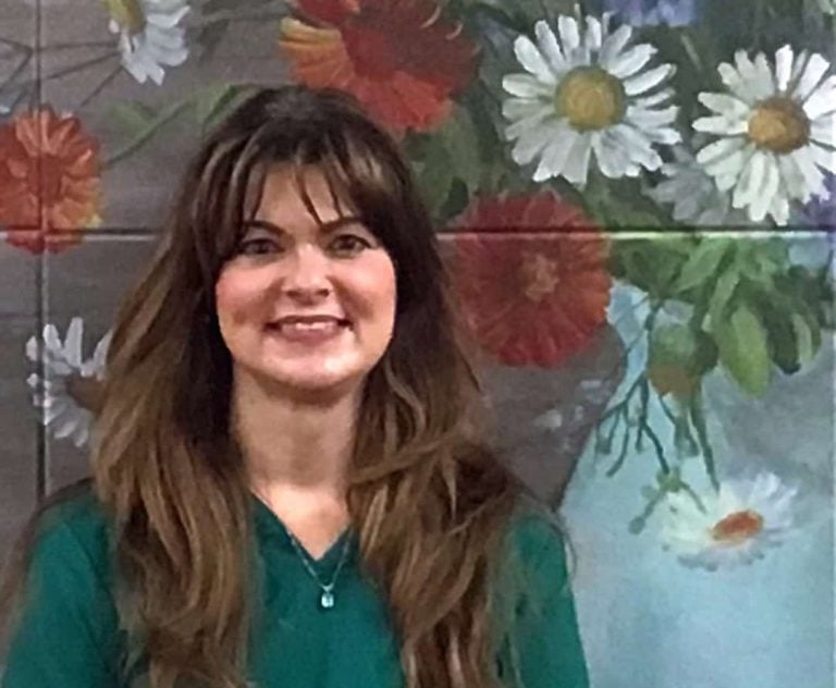 A woman with long brown hair and wispy bangs smiles in front of a painted flower mural.