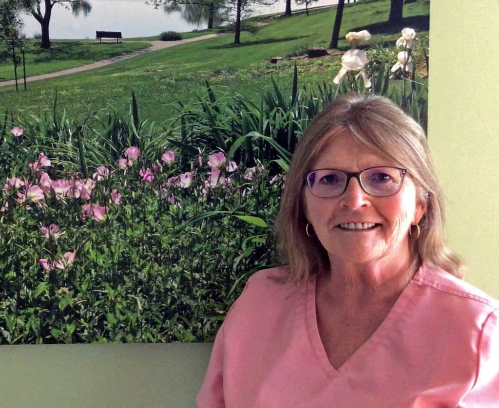 An older woman with shoulder length light brown and grey hair, smiling wearing glasses and gold hoop earrings. She is in pink scrubs standing in front of a blown up photo of a grassy green field with flowers and a lake in the distance.