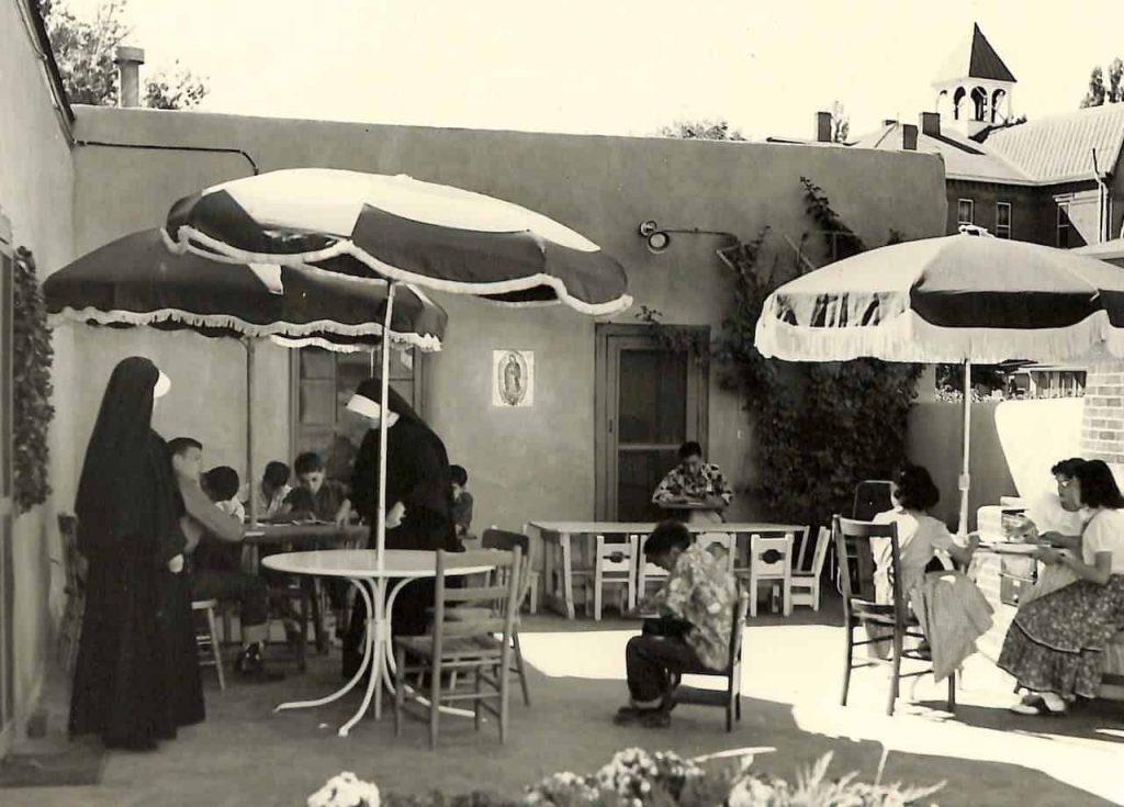 Students and teachers, nuns in habit, enjoy their time outside on a patio sitting in chairs underneath umbrellas.
