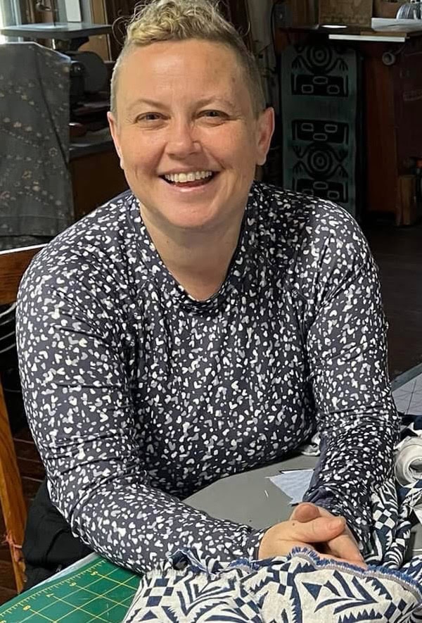 A middle aged white woman with short curly hair on top and shaved sides smiles in a studio. She is wearing. blue and white spotted long sleeve shirt.