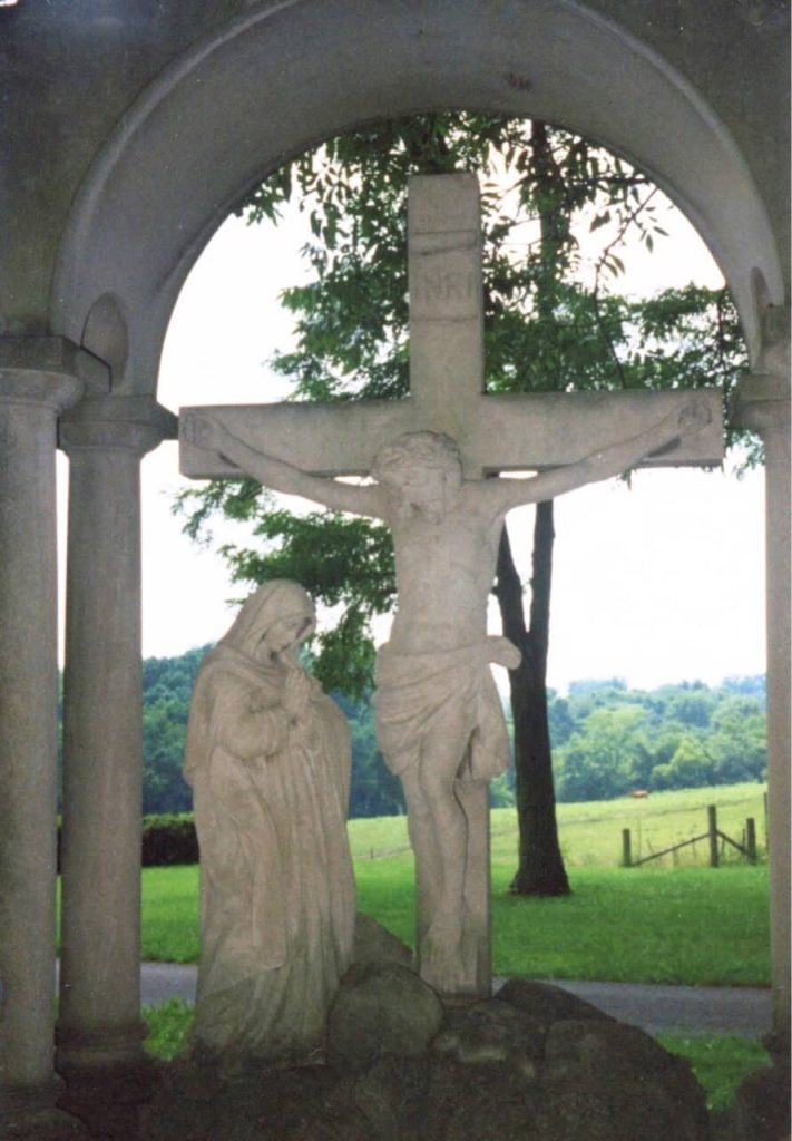A life-size sculpture of Mary praying at the foot of the cross where Jesus hangs. Green forest and fields are visible in the background.