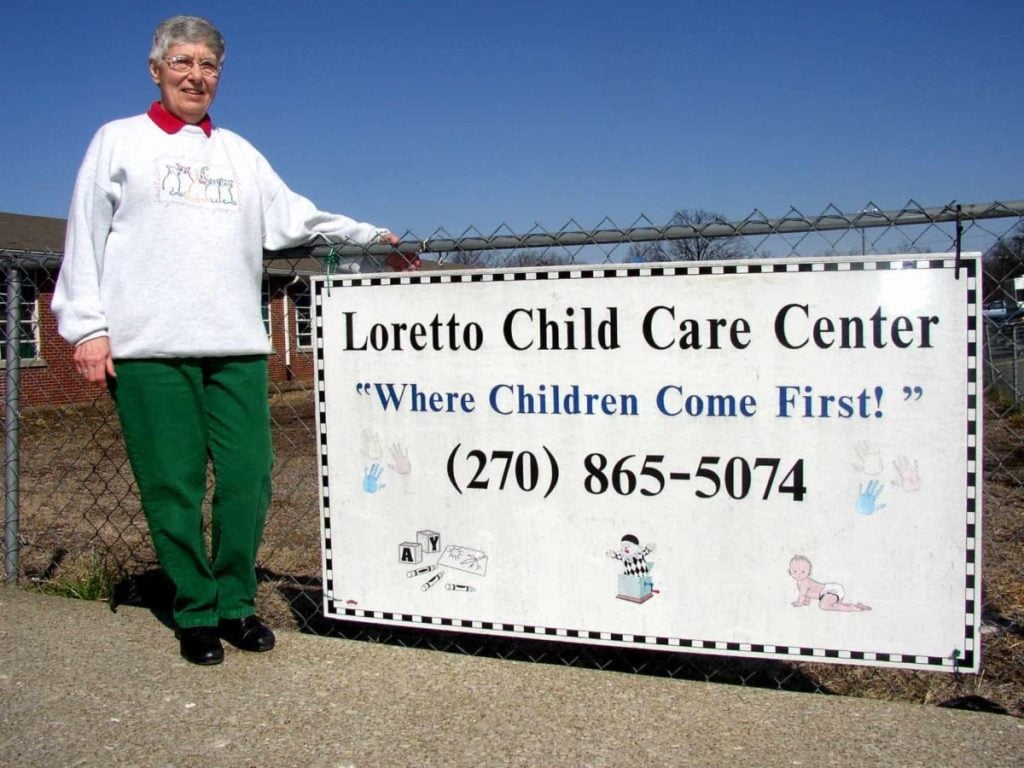 An older woman in green pants and a white sweatshirt smiling and standing next to a white sign outside that says "Loretto Child Care Center Where Children Come First"