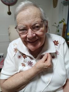 An elder white woman with short white hair wearing. white collared shirt with red flowers on it. Shes smiling and showing off a paperclip attached to the collar of her shirt.