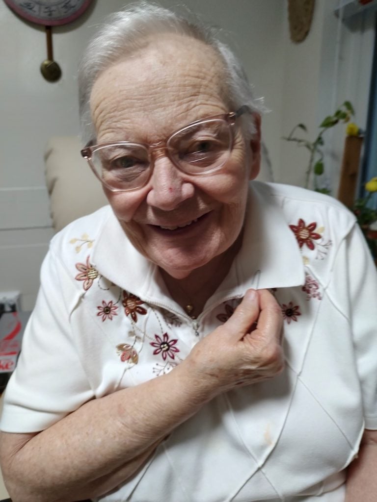 An elder white woman with short white hair wearing. white collared shirt with red flowers on it. Shes smiling and showing off a paperclip attached to the collar of her shirt.