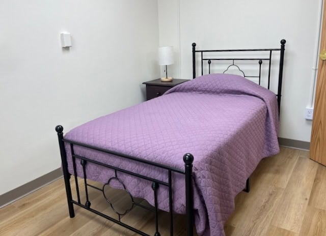 A bedroom with wood-finish floors, a single bed with a black metal bedframe and purple bedspread, and a bedside table with a reading lamp on it. The edge of the door can be seen on the right side of the photo.