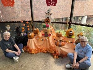 Two gray-haired women sit on the floor flanking a festive Halloween display of fall colors, foliage and pumpkins with sweet, painted jack o' lantern faces.