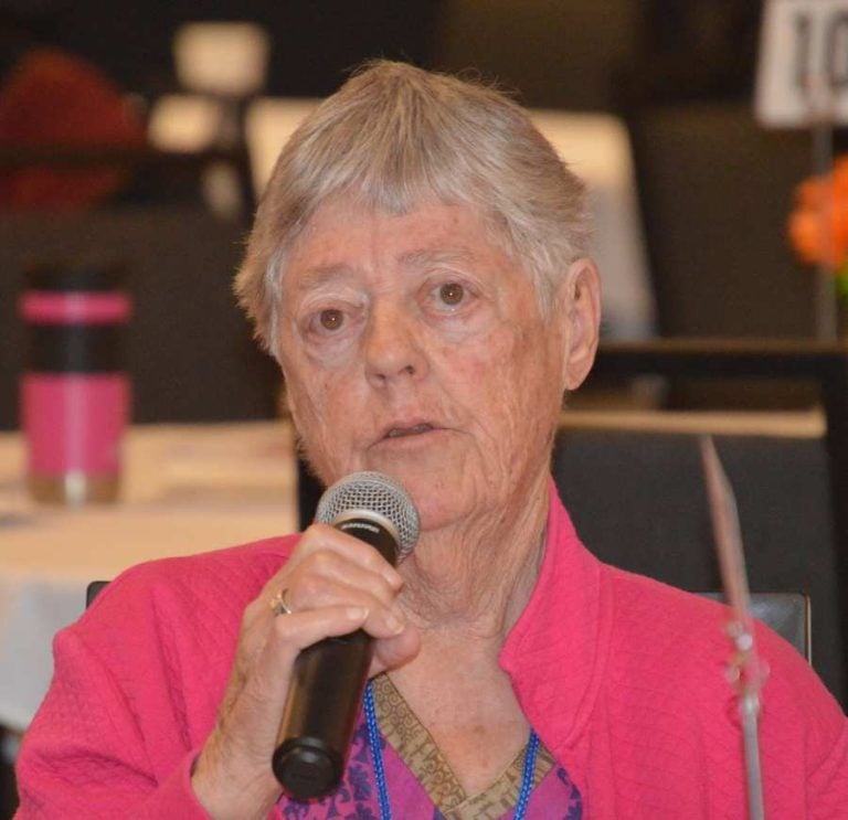 An older woman with short gray hair wearing a pink jacket and colorful shirt speaks into a microphone.