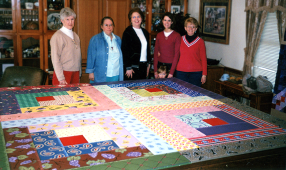 Five women and a child stand beside a large quilt laid out on a table.