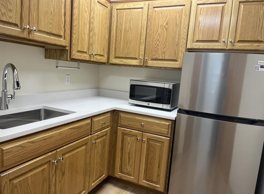 The corner of a kitchen with wood-finish cabinets, stainless steel fridge and small microwave and a double-wide sink.