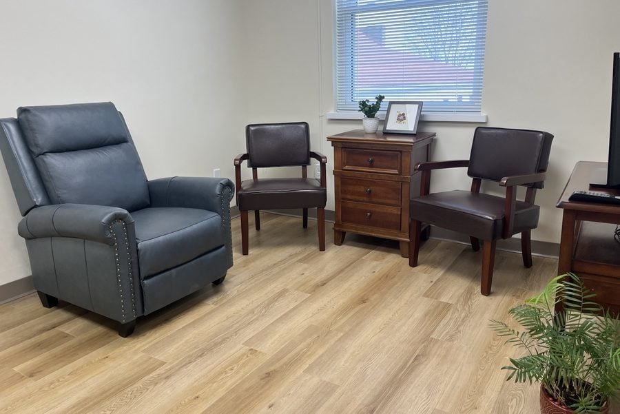 A comfortable living room space with a large window and wood-finish floor, furnished with a recliner, two additional chairs and a tv table with a tv. A small fern is on the floor next to the tv table.