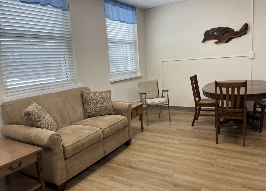 A comfortable living space list by two large windows and displaying wood-finish floors, a comfortable sofa, and a round, expandable dining table with chairs.