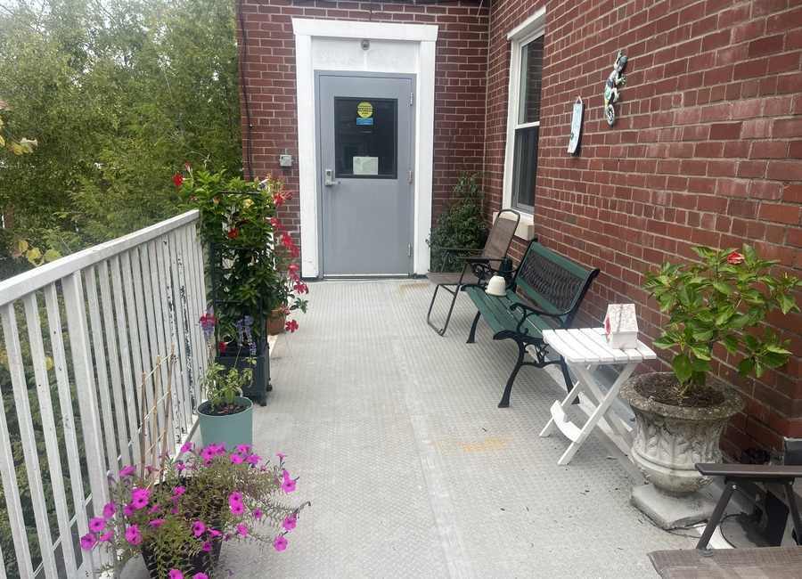An outdoor patio formed by a white railing on the left and a brick building on the right. Patio furniture and potted flowers give it a welcoming look.