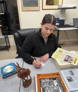 A dark-haired woman writing notes sits at a desk with CDs, publications and a gazelle carving.