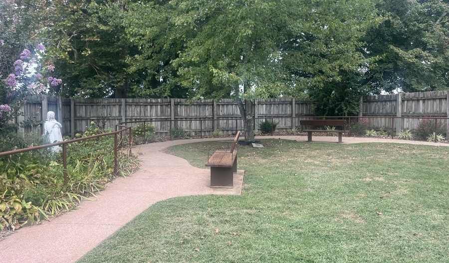 A handrail-lined path winds through a private green space which is protected by a wooden privacy fence. A white statue stands in the greenery to the side of the path, while two benches are placed at intervals along the path.