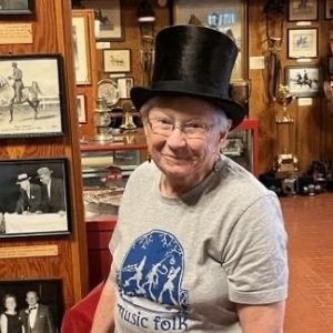 Older woman wearing a "Music Folk" t-shirt and top hat stands in a display of old photographs.