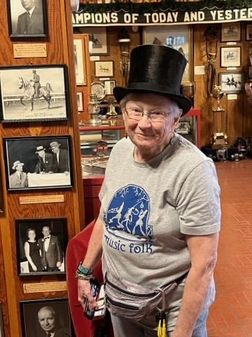 Older woman wearing a "Music Folk" t-shirt and top hat stands amidst a display of historic photos of horses and their trainers.